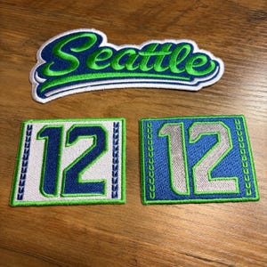 May include: Embroidered patches featuring the word "Seattle" in green and blue script, and two patches with the number "12". The patches have a white border and are on a wooden surface.