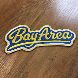 May include: Embroidered patch with the words "Bay Area" in a stylized script. The text is yellow with a blue outline and a white border. The patch is on a wooden surface.