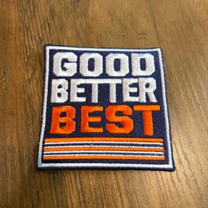 May include: A square embroidered patch with a dark blue background and a white border. The text GOOD BETTER is stitched in white, while the word BEST is in orange. Three horizontal stripes in orange and white appear at the bottom of the patch.