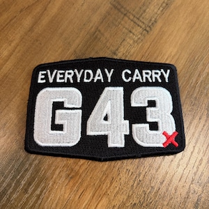 May include: A rectangular black embroidered patch with a black border. The text "EVERYDAY CARRY" is stitched in white at the top. Below, the large white alphanumeric characters "G43" are displayed, with a small red "X" stitched at the bottom right.