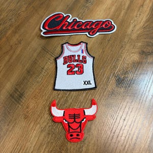 May include: Three embroidered patches featuring Chicago Bulls basketball team branding. One patch displays the word Chicago in red script, one is a white jersey with Bulls 23 and XXL text, and one is a red and white bull head logo emblem.