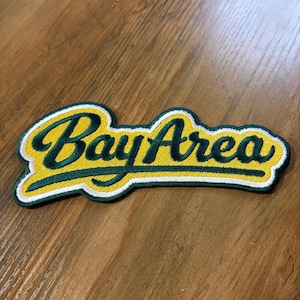 May include: Embroidered patch with the words "Bay Area" in a script font. The text is green with a yellow background and white border. The patch is rectangular with rounded edges.
