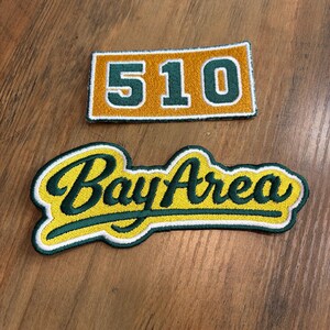 May include: Two embroidered patches. One rectangular patch features the number 510 in dark green on a gold background. The second patch features the text Bay Area in dark green cursive script on a yellow background with a dark green border.
