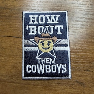 May include: Embroidered patch with the text "HOW 'BOUT THEM COWBOYS" in white lettering on a navy blue background. The patch features a smiling cowboy emoji wearing a brown cowboy hat, set within a white star.