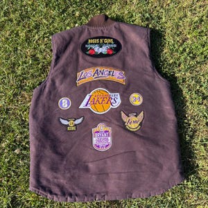 May include: A brown vest with multiple patches, including a "Roses N' Guns" patch, a "Los Angeles Lakers" patch, and a basketball patch. Other patches include the numbers 8 and 24, and the name "Kobe".