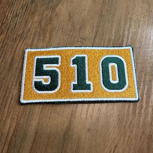 May include: A rectangular embroidered patch featuring the number 510 in dark green with a white outline, set against a vibrant gold background. The patch has a dark green stitched border, creating a clean and defined edge for the numeric design.