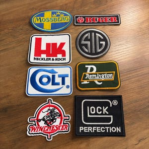 May include: A collection of embroidered patches featuring various brand logos, including Mossberg, Ruger, Heckler & Koch, SIG, Colt, Remington, Winchester, and Glock. The patches are in various shapes and colors, with detailed stitching.