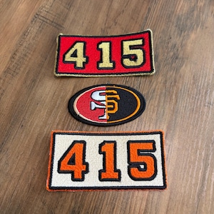 May include: Three embroidered patches. The top rectangular patch is red with gold numbers 415 and a black border. The middle oval patch features a split red and black design with a white and gold logo. The bottom rectangular patch is white with orange numbers 415 and a black border.