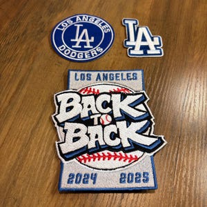 May include: Three embroidered patches featuring Los Angeles Dodgers branding. One circular blue patch with white text, one blue and white LA logo patch, and one rectangular patch with a baseball graphic, the text BACK TO BACK, and 2024 2025.