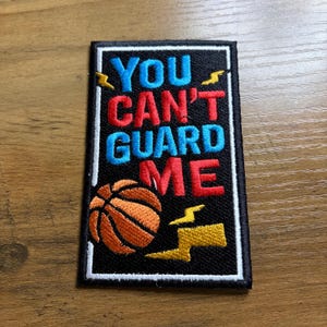 May include: Embroidered patch with the text "YOU CAN'T GUARD ME" in blue and red, with a basketball and yellow lightning bolts on a black background. The patch has a white border.