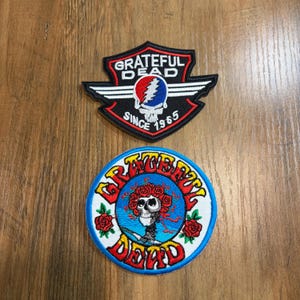May include: Two embroidered patches. The top patch is a black shield shape with white wings, a skull icon, and the text "GRATEFUL DEAD SINCE 1965". The bottom patch is circular with a blue center, a skull wearing a crown of red roses, and the text "GRATEFUL DEAD" in red.