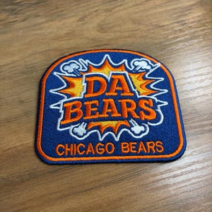 May include: Embroidered patch with a blue background and orange and white accents. The patch features the words "DA BEARS" in orange, with "CHICAGO BEARS" in white below. The design includes a burst of orange and white lightning bolts.