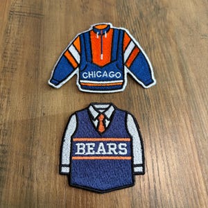 May include: Two embroidered patches shaped like clothing. The top patch is a blue and orange pullover with white stripes and the word CHICAGO. The bottom patch is a blue vest over a white collared shirt with an orange tie and the word BEARS.