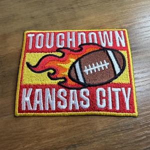May include: Embroidered patch featuring a brown football with white stripes and a flame design. The patch has the words "TOUCHDOWN" and "KANSAS CITY" in white letters on a red and yellow background.