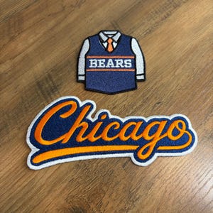 May include: Two embroidered patches. One patch is a navy blue vest over a white shirt with an orange tie and the word BEARS in white text. The second patch features the word Chicago in orange cursive script with a navy blue outline and white border.