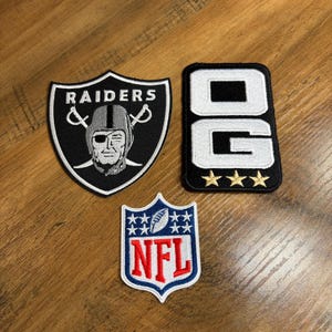 May include: Three embroidered patches: a black and white shield-shaped Raiders logo, a rectangular black patch with white letters OG and three gold stars, and a shield-shaped NFL logo with red letters, a white football, and blue accents.