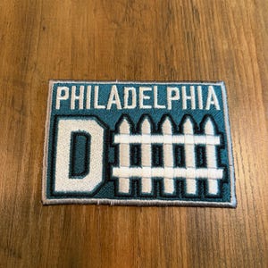 May include: A rectangular embroidered patch with a teal background and a light gray border. The text PHILADELPHIA is stitched in white at the top. Below, a large white letter D sits next to a white picket fence graphic, both outlined in dark blue.