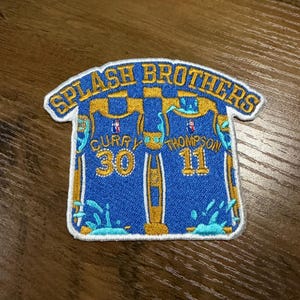 May include: Embroidered patch featuring a blue and gold basketball jersey design. The patch reads "SPLASH BROTHERS" at the top and includes the numbers 30 and 11, with the names "CURRY" and "THOMPSON" below. The patch has a white border.