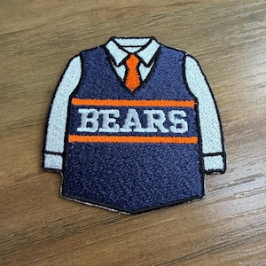 May include: Embroidered patch featuring a navy blue sweater vest design with the word "BEARS" in white block letters. The vest has an orange stripe and a white collared shirt with an orange tie. The patch is likely for clothing or accessories.