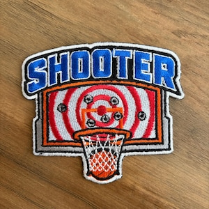 May include: Embroidered patch featuring a basketball hoop and backboard with a target design. The word "SHOOTER" is in blue letters with a white and orange outline. The patch has a white border.