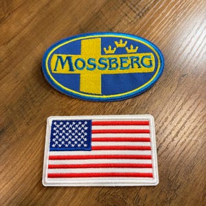 May include: Two embroidered patches: one oval blue patch with a yellow cross and the word "MOSSBERG", and a rectangular patch featuring the American flag with red and white stripes and a blue field of stars.