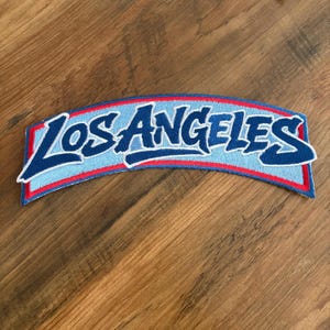 May include: Embroidered patch with the text "LOS ANGELES" in a stylized, cursive font. The text is in dark blue with a white outline, set against a light blue background. The patch has a red border and a white edge.