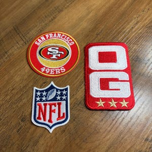 May include: Three embroidered patches including a circular San Francisco 49ers logo with red, gold, and white, a shield-shaped NFL logo in blue, red, and white, and a rectangular red patch with white letters OG and three gold stars at the bottom.