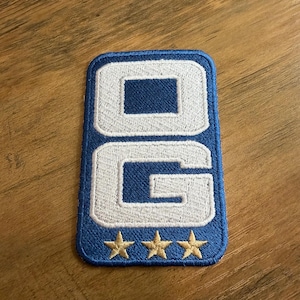 May include: A rectangular embroidered patch with a blue border and background. The patch features the white letters O and G stacked vertically. Below the letters are three small, gold-colored embroidered stars arranged in a horizontal row.
