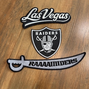 May include: Three embroidered patches featuring a black and white Las Vegas script logo, a Raiders shield emblem with a central pirate figure wearing an eye patch, and a curved sword-shaped patch with the text RAAAAIIDERS in black lettering.