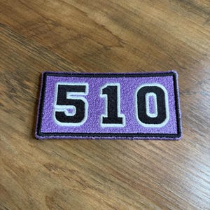 May include: A rectangular embroidered patch featuring the number 510 in black with a white outline, set against a lavender purple background. The patch has a thick black border that frames the entire design, creating a clean and defined edge.