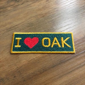 May include: Embroidered patch with the words "I ❤️ OAK" in yellow letters and a red heart on a dark green background. The patch has a yellow border and is rectangular in shape.