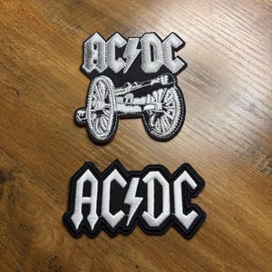 May include: Two embroidered patches featuring the band AC/DC. One patch shows the band's logo with a lightning bolt, the other features the logo above a detailed cannon design. Both patches have a black background with white stitching.