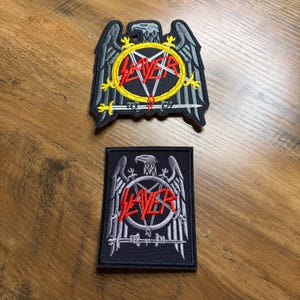 May include: Two Slayer patches. The top patch is an eagle design with the word "SLAYER" in red inside a yellow circle. The bottom patch is a rectangular design with the same logo. Both patches are black with gray and red accents.