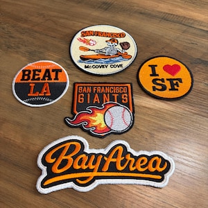 May include: A collection of embroidered patches with baseball themes. Designs include "Beat LA" with a baseball, "I ❤️ SF", "San Francisco Giants" with a baseball and flames, a "Bay Area" patch, and a patch with a skeleton kayaking.