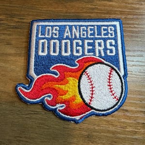 May include: Embroidered patch featuring the Los Angeles Dodgers logo. The patch has a blue background with white text that reads "LOS ANGELES DODGERS." A baseball with red stitching is surrounded by flames in shades of red, orange, and yellow.