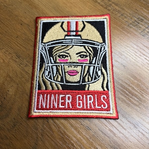 May include: Embroidered patch featuring a woman in a football helmet. The patch has a black background with a gold helmet and the text "NINER GIRLS" in red. The woman has pink eye makeup and a pink lip color.