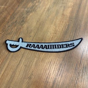 May include: A curved sword-shaped embroidered patch with a black border and light blue fill. The text RAAAAIIDERS is stitched in black across the center of the blade. The hilt features a circular guard and a pommel, all outlined in black thread.