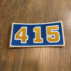 May include: A rectangular embroidered patch featuring the number 415 in gold thread with a white outline, set against a solid blue background. The patch is finished with a gold-colored stitched border, creating a clean and defined edge.