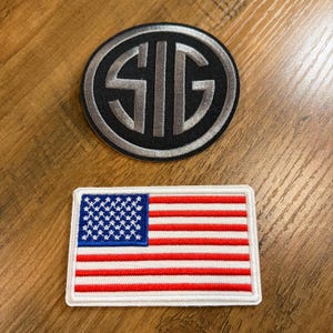 May include: Two embroidered patches on a wooden surface. One is a black and silver circular patch with the letters "SIG". The other is a rectangular patch of the American flag with red and white stripes and a blue field of stars.