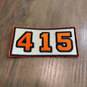 May include: A rectangular embroidered patch featuring the number 415 in bold orange digits with a thick black outline. The background of the patch is a light cream color, and the entire design is bordered by a thin orange stitched edge.