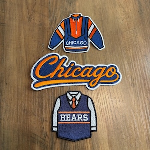 May include: Three embroidered patches featuring Chicago sports themes. The top patch is a blue and orange pullover with the word Chicago. The middle patch is the word Chicago in orange script with a blue outline. The bottom patch is a blue vest.