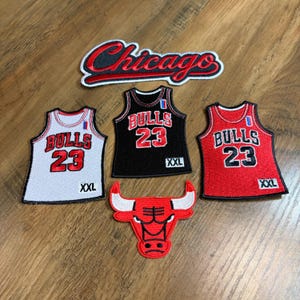 May include: A collection of embroidered patches featuring a Chicago script logo, a red bull head logo, and three basketball jerseys in white, black, and red. Each jersey displays the text Bulls 23 and XXL, with a small blue and red accent on the shoulder.