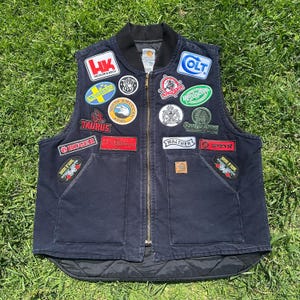 Op de afbeelding: Een marineblauwe Carhartt-vest met een zwarte kraag en talrijke patches. Patches met logo's van vuurwapenfabrikanten zoals Colt, Walther en Taurus. Het vest heeft een ritssluiting aan de voorkant en twee voorzakken, tegen een achtergrond van groen gras.