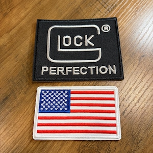 May include: Two embroidered patches. One is black with the word "GLOCK" in white, and the word "PERFECTION" below. The other is a rectangular patch of the American flag with red and white stripes and a blue field of white stars.