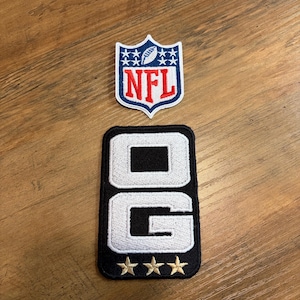 May include: A shield-shaped NFL logo patch featuring a football, stars, and red lettering on a blue and white background, positioned above a rectangular black patch with white embroidered letters OG and three gold stars at the bottom edge.