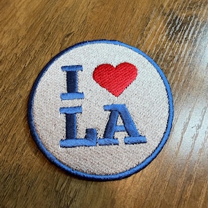 May include: Embroidered patch with the text "I ❤️ LA" in blue, with a red heart replacing the word "love". The patch is circular, with a light tan background and a blue border. The patch is likely for clothing or accessories.