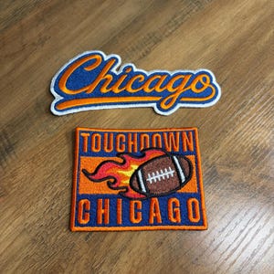 May include: Two embroidered patches. The top patch features the word Chicago in orange script with a blue outline. The bottom rectangular patch features the text Touchdown Chicago in orange block letters with a brown football graphic on fire.