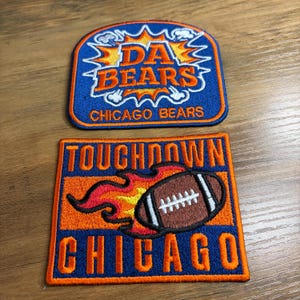 May include: Two embroidered patches featuring the Chicago Bears football team. One patch reads "DA BEARS" in orange lettering on a blue background. The other patch has a football with flames and the word "TOUCHDOWN" and "CHICAGO".