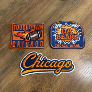 May include: Three embroidered patches featuring Chicago Bears themes. One rectangular patch displays a football with flames and the text TOUGHDOWN CHICAGO. A second shield-shaped patch features the text DA BEARS CHICAGO BEARS. A third script patch says Chicago.