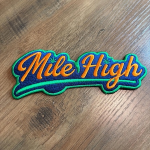 May include: An embroidered patch featuring the text Mile High in orange cursive script with a dark blue background and a bright green border. Small green leaf icons are integrated into the lettering above the letters i and h.